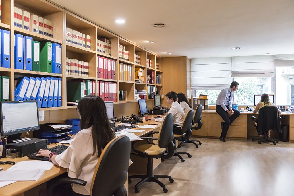 Varias personas trabajando con ordenadores y documentación en un despacho profesional con estanterías llenas de archivadores y libros jurídicos.