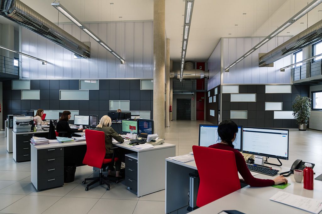Varias personas trabajando con ordenadores en una oficina administrativa amplia, integrada en un edificio de estilo industrial con techos altos y conductos visibles.