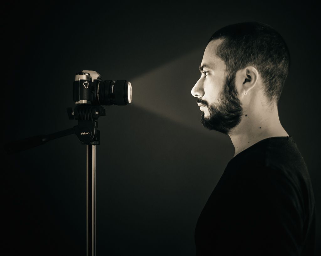 Retrato de fotógrafo junto a una cámara sobre trípode en un estudio, con iluminación dirigida y fondo oscuro.