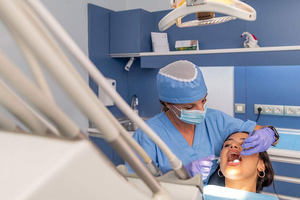Profesional sanitaria realizando un tratamiento odontológico a una paciente en una consulta dental, con instrumental clínico y lámpara de exploración.