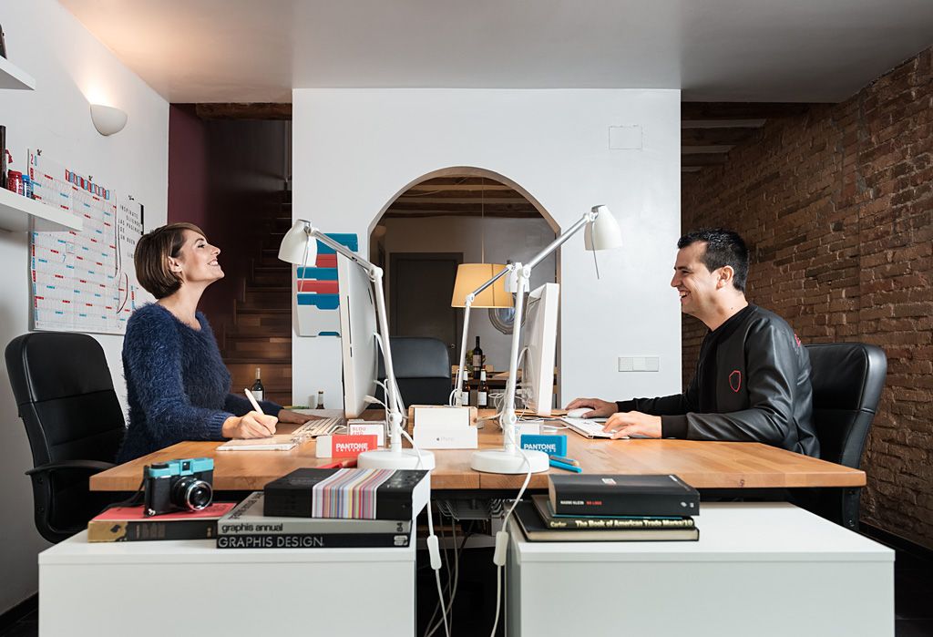 Dos personas trabajando frente a ordenadores en un estudio creativo, con muestras de color, libros de diseño y material de trabajo sobre la mesa.