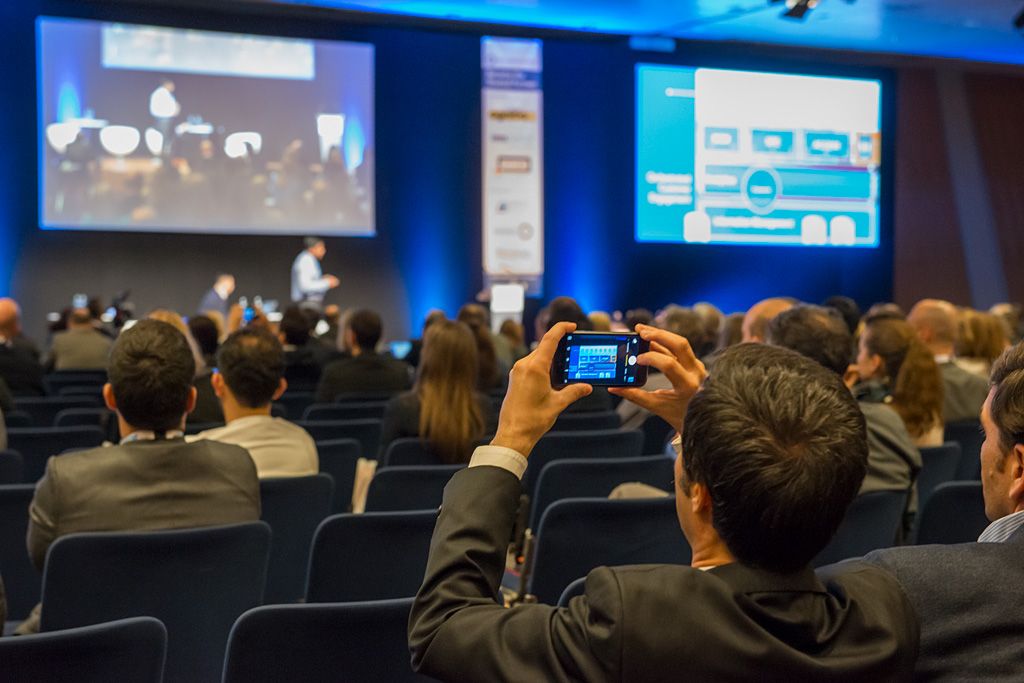 Asistentes en un evento corporativo durante una ponencia, con una persona grabando la presentación con el móvil.