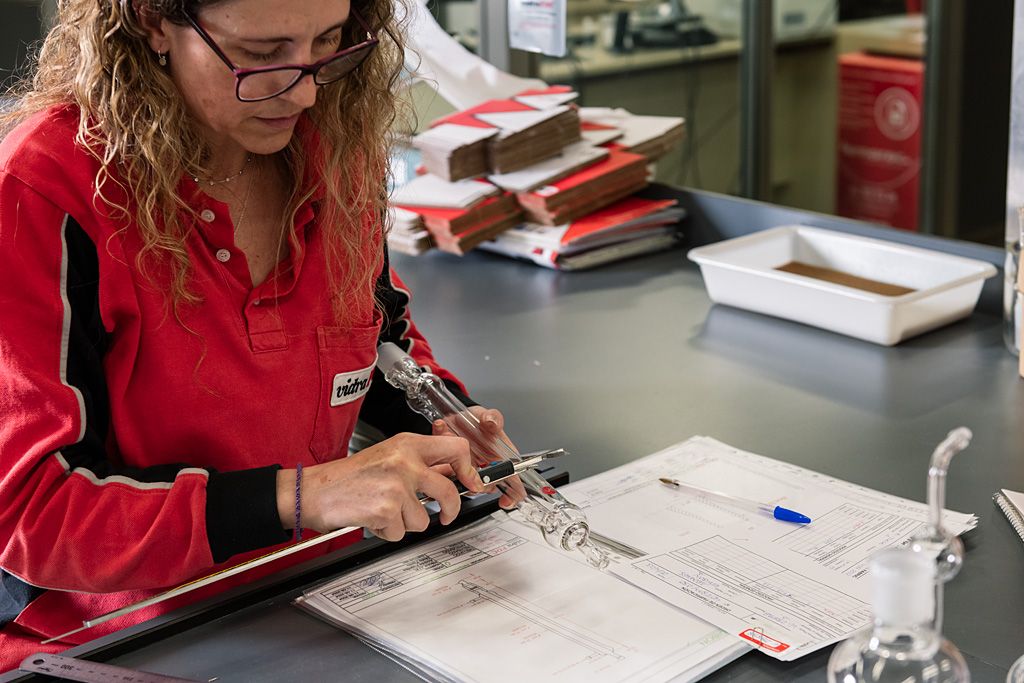 Profesional midiendo una pieza de vidrio técnico sobre documentación de fabricación.