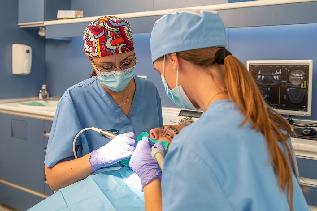 Dos profesionales sanitarias realizando un tratamiento dental a un paciente en una clínica odontológica, con material médico y pantalla de diagnóstico al fondo.